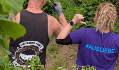 Zwei Personen von hinten bei einer ehrenamtlichen Aktion im Grünen. Links ein Mann mit Goitzsche Front Chaos Crew Tanktop, rechts eine Frau mit violettem T-Shirt mit der Aufschrift #AUSLIEBE – beide zeigen mit dem Daumen auf sich.