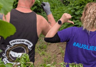 Zwei Personen von hinten bei einer ehrenamtlichen Aktion im Grünen. Links ein Mann mit Goitzsche Front Chaos Crew Tanktop, rechts eine Frau mit violettem T-Shirt mit der Aufschrift #AUSLIEBE – beide zeigen mit dem Daumen auf sich.